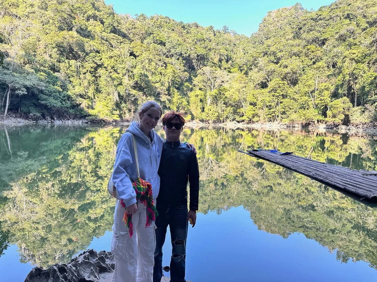 Two people stand by a serene lake, surrounded by lush green trees. One holds a colorful cloth. A bamboo raft is visible on the water.