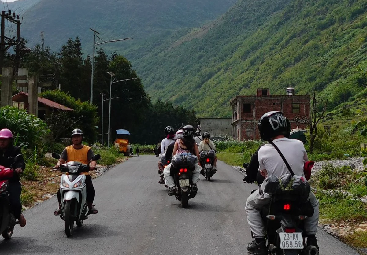 Motorcyclists ride on a rural road flanked by greenery and mountains. One wears a pink helmet, another a yellow vest. A building is in the background.