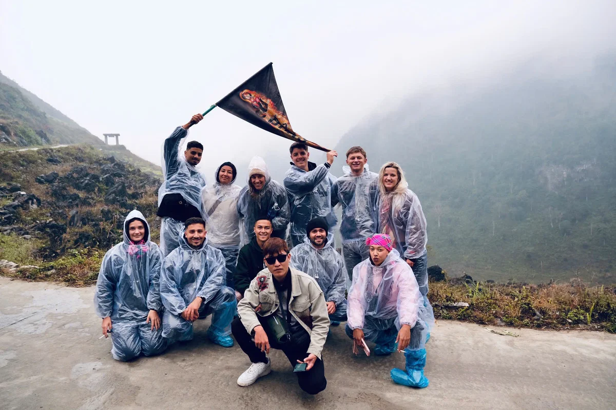 Group of people in rain ponchos pose on a foggy mountain path. One holds a flag. Mood is cheerful and adventurous. Sparse greenery.