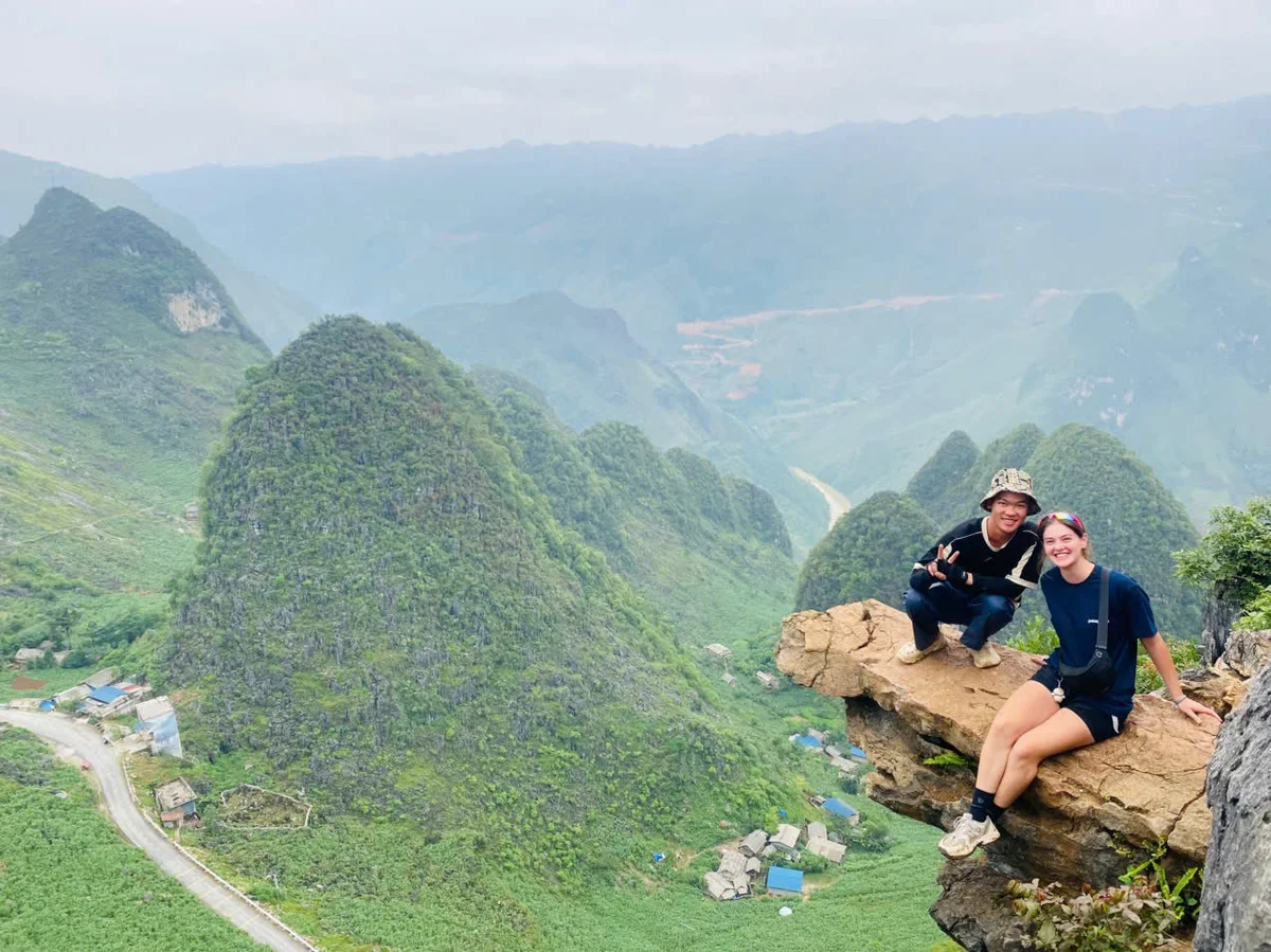 Two people on a cliff edge, smiling with peace signs. Lush green mountains in the background, overcast sky, creating a serene mood.