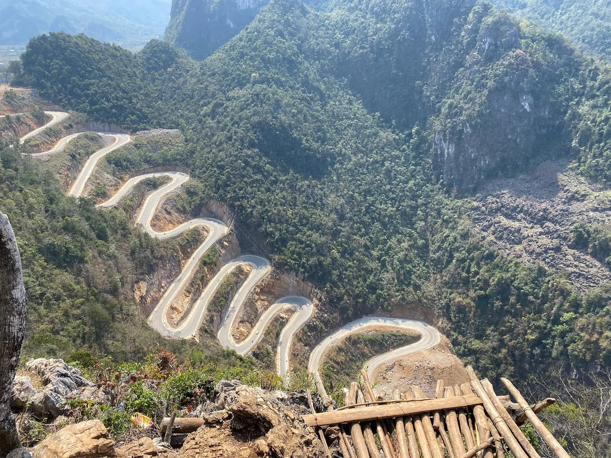A winding mountain road with sharp curves snakes through lush green hills. Foreground shows wooden logs and rocky terrain under a clear sky.
