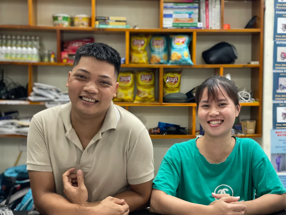 Two people smiling in a shop with shelves of snacks and books in the background. They appear cheerful and relaxed.
