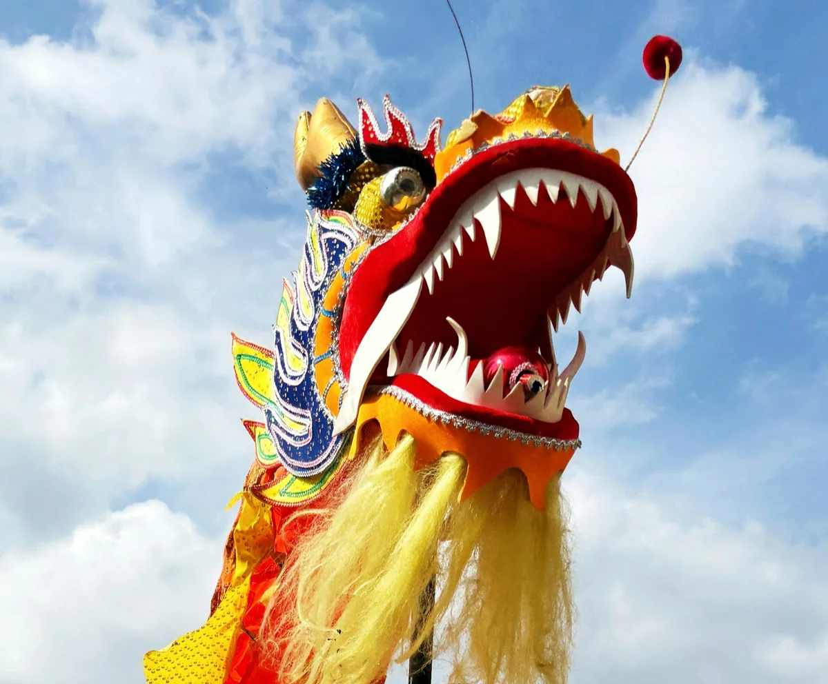 Colorful Chinese dragon head with wide open mouth against a blue sky. Bright red, yellow, and blue patterns create a festive mood.