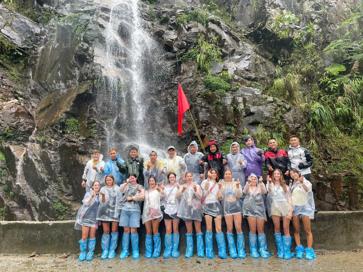 Group posing in rain ponchos and blue shoe covers by a waterfall and rocks, holding a red flag, with expressions of joy and adventure.