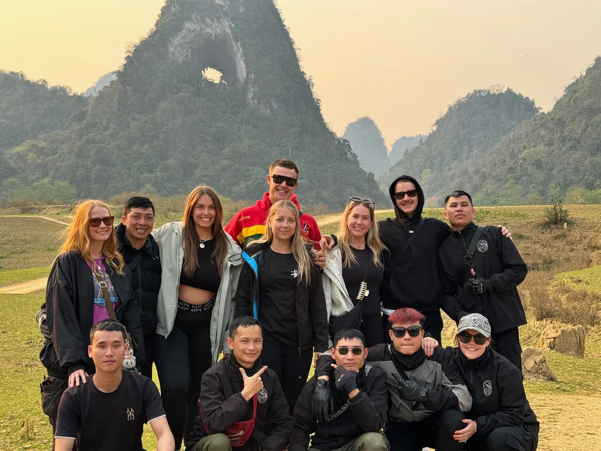 A group of people posing together outdoors with lush green hills and a large, round rock formation in the background under a clear sky.