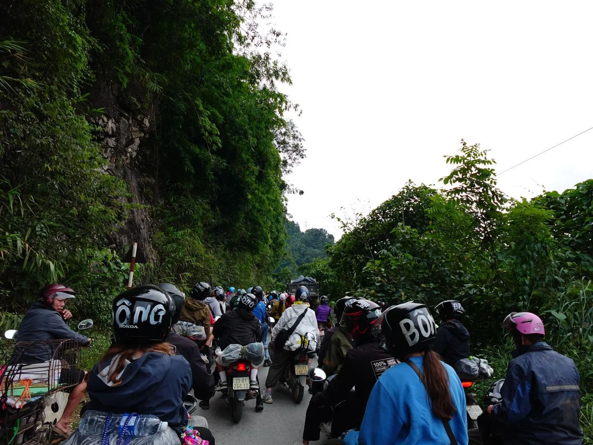 Motorcyclists with helmets ride on a narrow road surrounded by lush greenery. The atmosphere is crowded and lively.