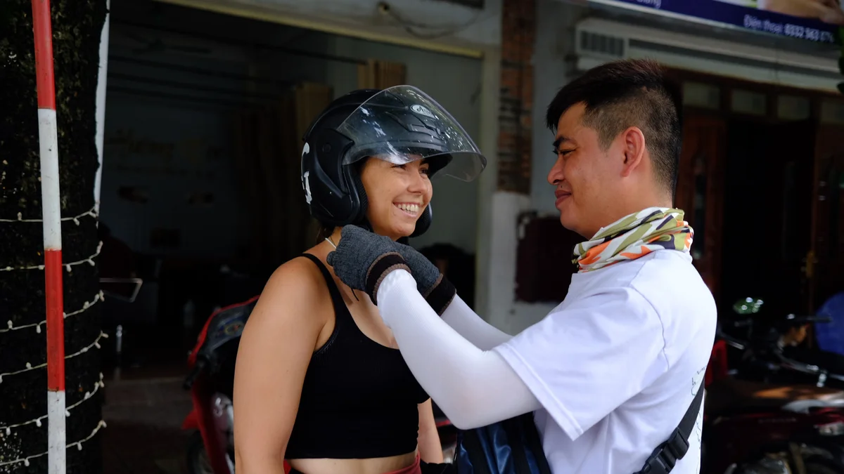 Man helps woman adjust helmet, both smiling. Woman wears black top; background shows a shop with a scooter and blurred sign.