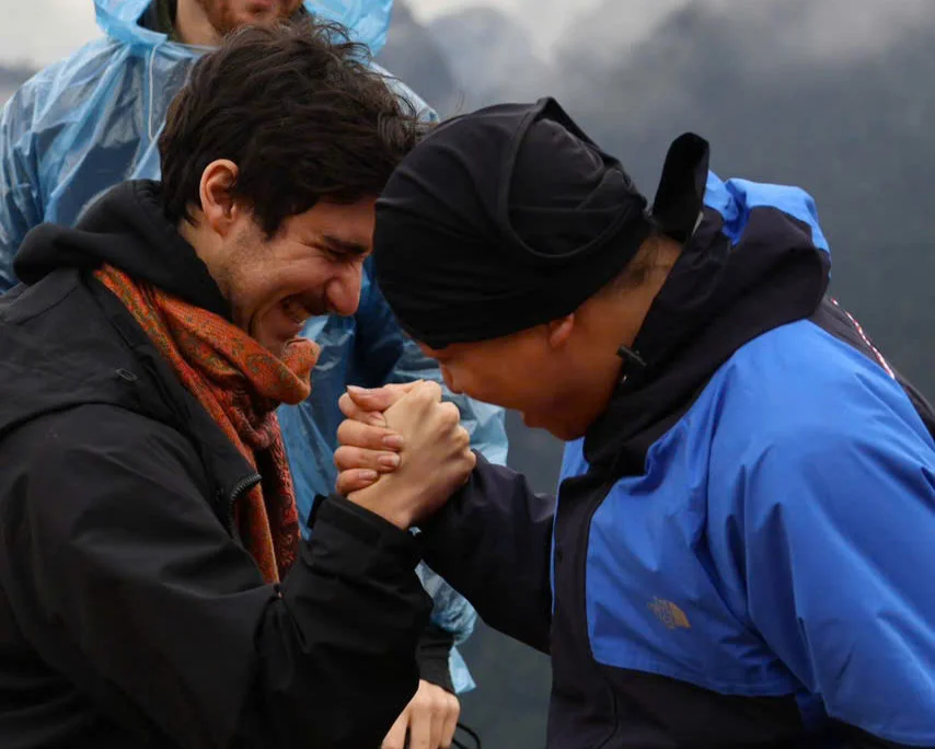 Two men happily arm wrestling outdoors, one in a black jacket with an orange scarf, the other in a blue raincoat, cloudy mountains behind.