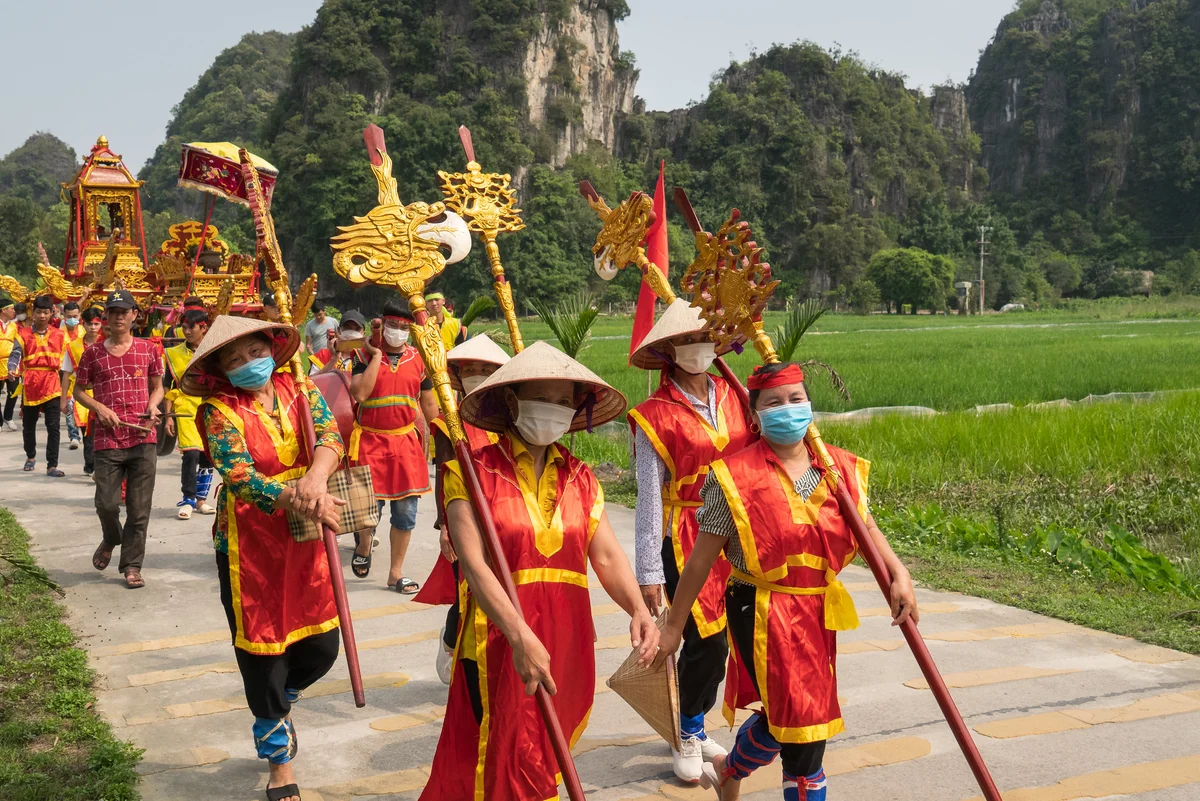 Procession of people in red and yellow robes carrying ornate staffs, walking on a path alongside a green field with limestone cliffs behind.