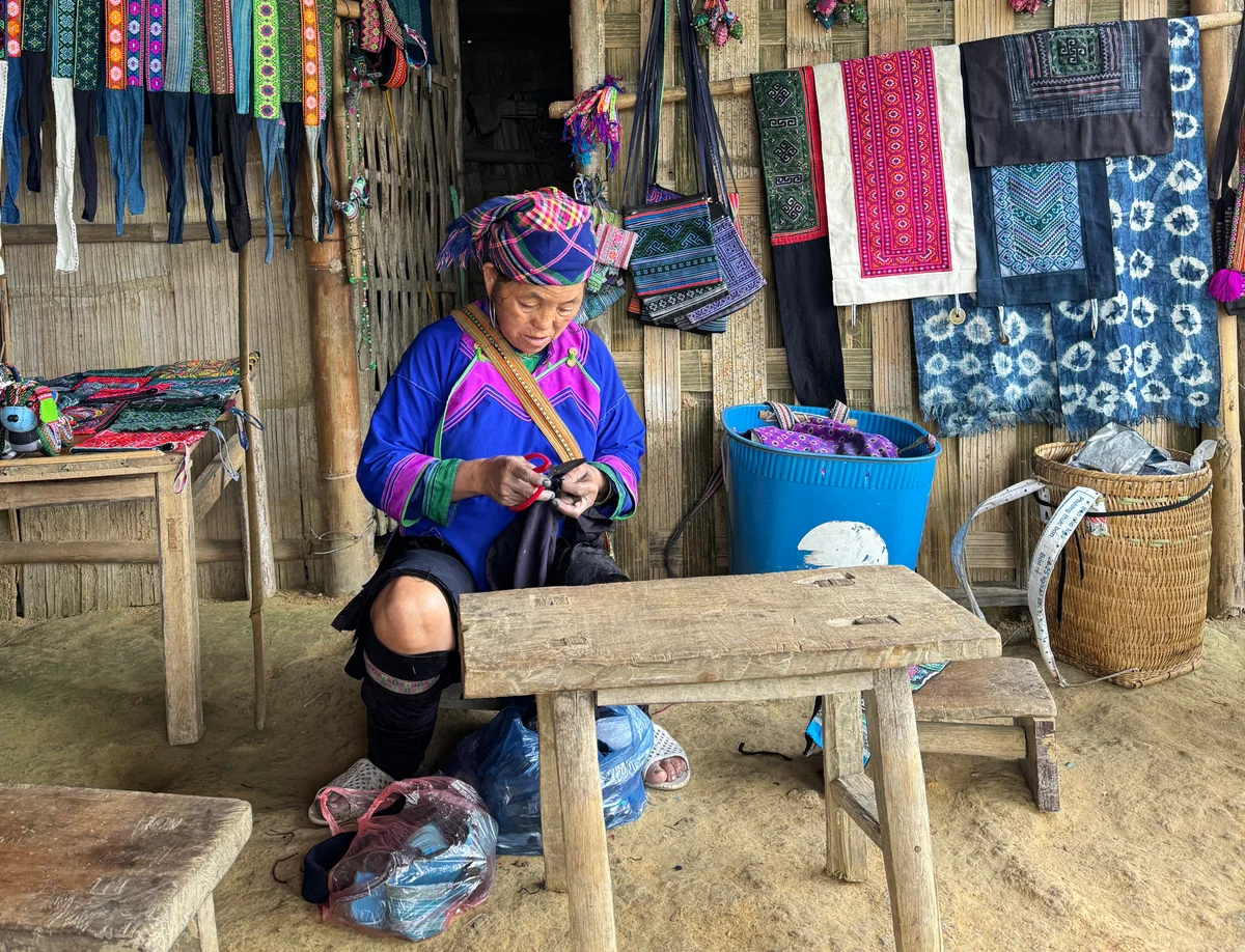 Woman in vibrant traditional attire sewing in a rustic shop. Colorful fabrics hang around, with wooden tables and baskets nearby.