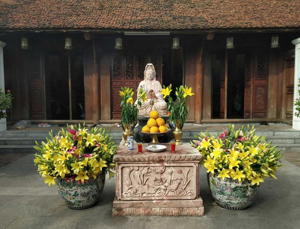 Stone statue on ornate altar with yellow lilies and fruit offering in front of a traditional wooden temple. Peaceful and serene setting.