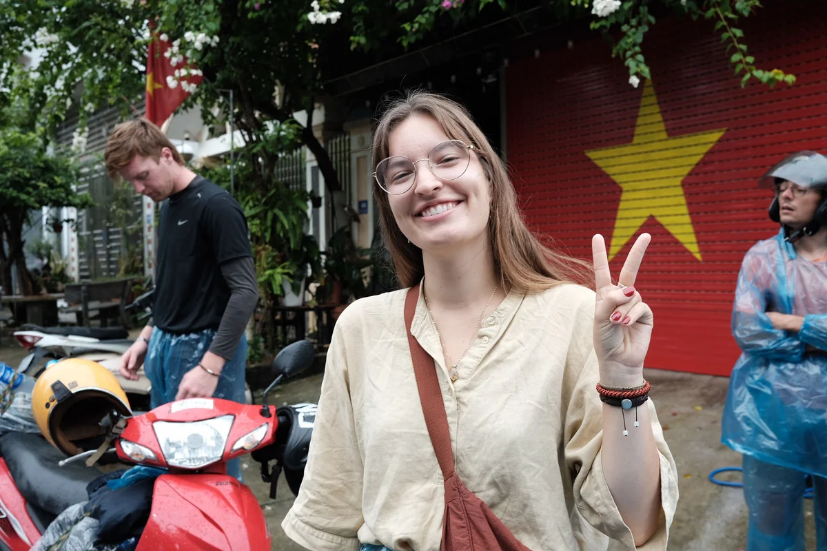 Woman smiling and flashing a peace sign in front of a red wall with a yellow star. Scooters and two people in the background. Casual mood.