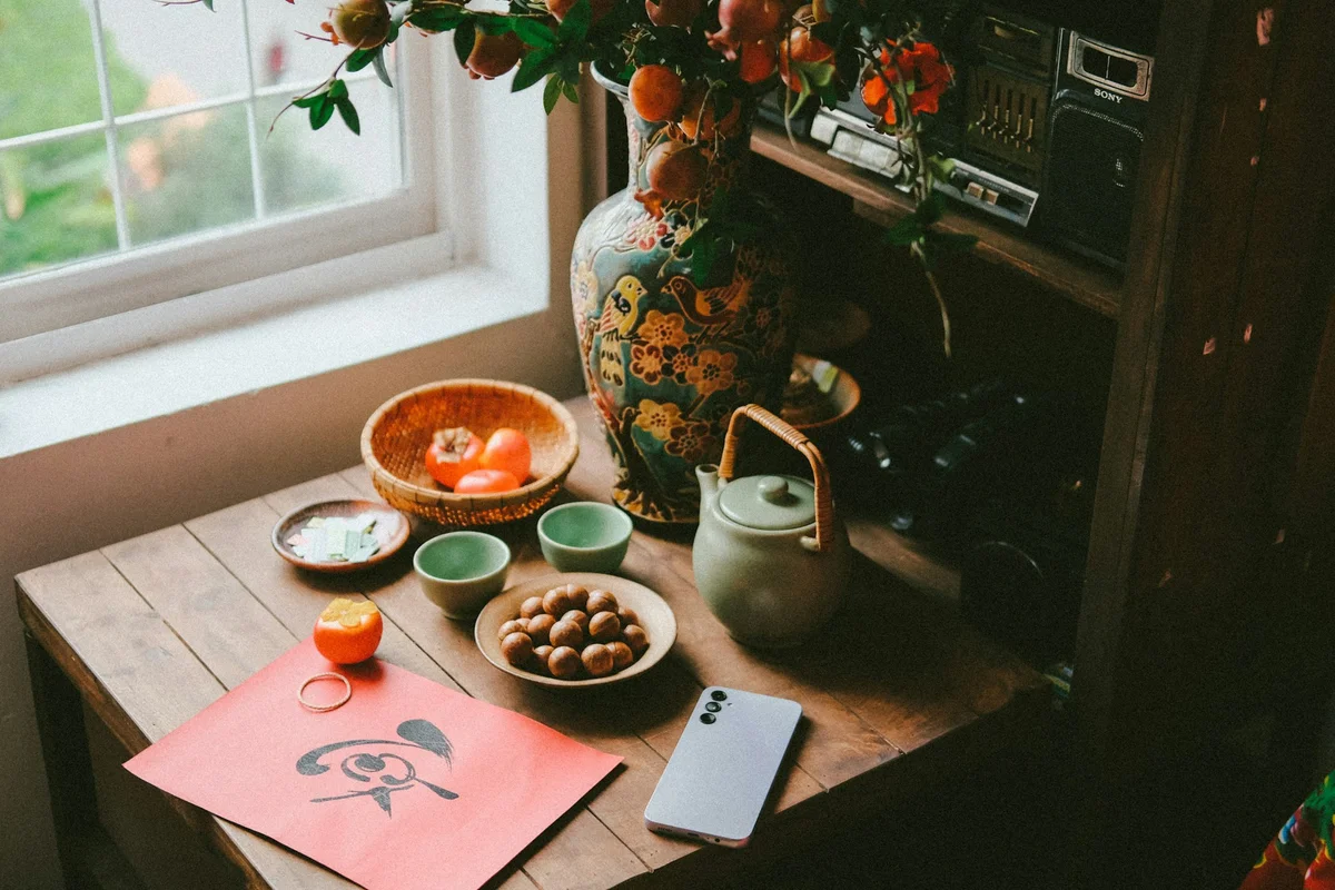 Decorative table with a floral vase, teapot, red paper with artwork, fruits, cups, and smartphone. Sunlight through window, cozy mood.