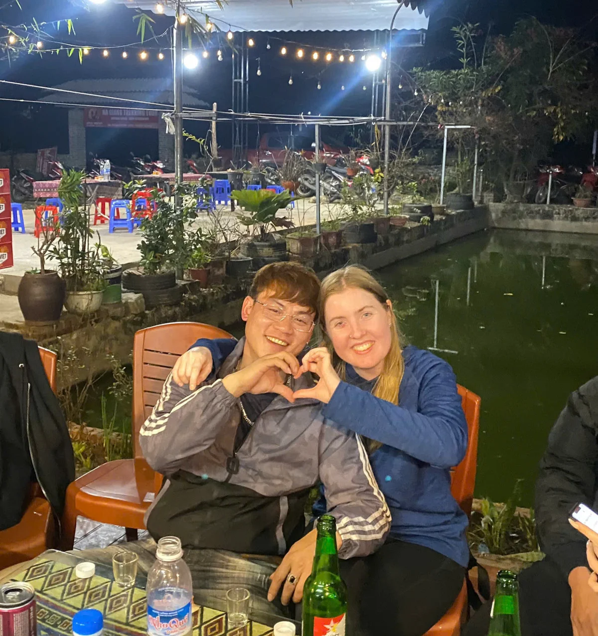Two people are sitting outdoors, smiling, and forming a heart shape with their hands. Background has string lights and a pond. Night setting.
