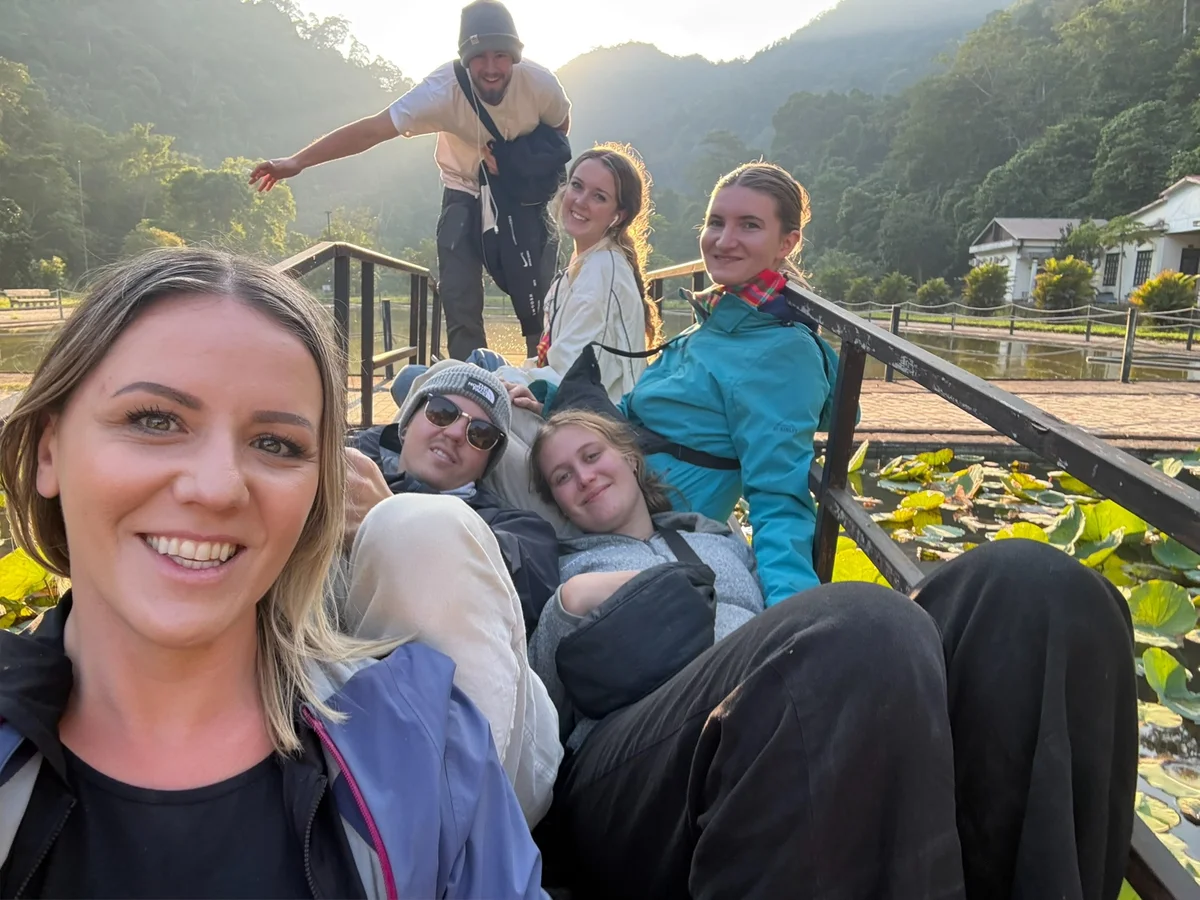 Group of friends smiling on a bridge with lush green hills and a pond in the background. Warm sunlight; relaxed, joyful mood.