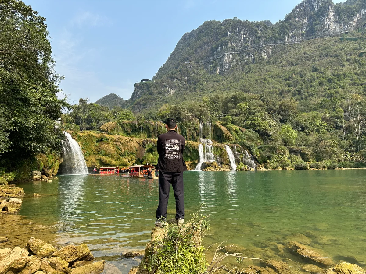 A man in a black jacket and trousers poses in front of a waterfall