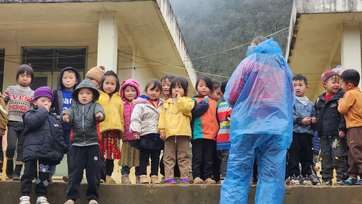 Children in colorful jackets stand on a concrete step, facing a person in a blue raincoat. Overcast day, rural building in background.