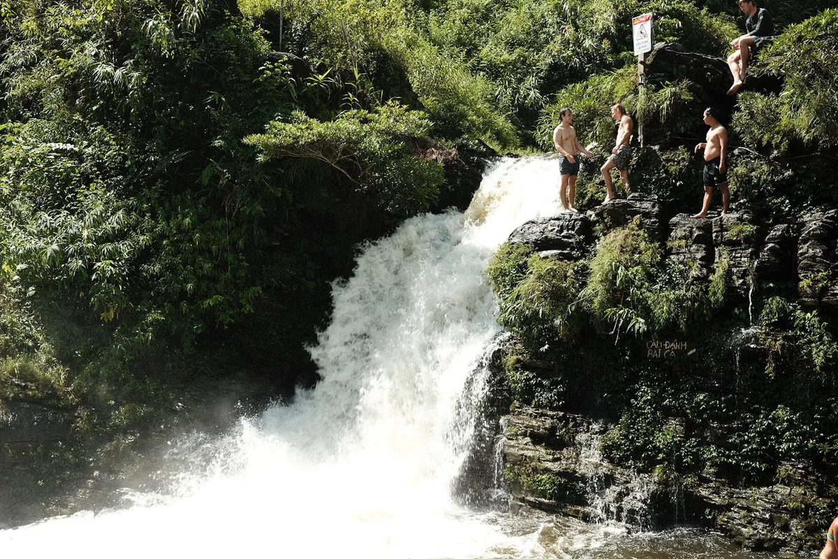 Four people stand on a rocky ledge by a waterfall in a lush green forest. A "Danger" sign is visible above the waterfall.