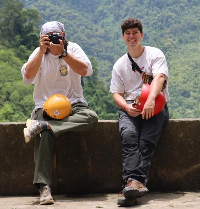Two people smile at the camera with mountains behind, one is aiming a camera at the person taking the photo