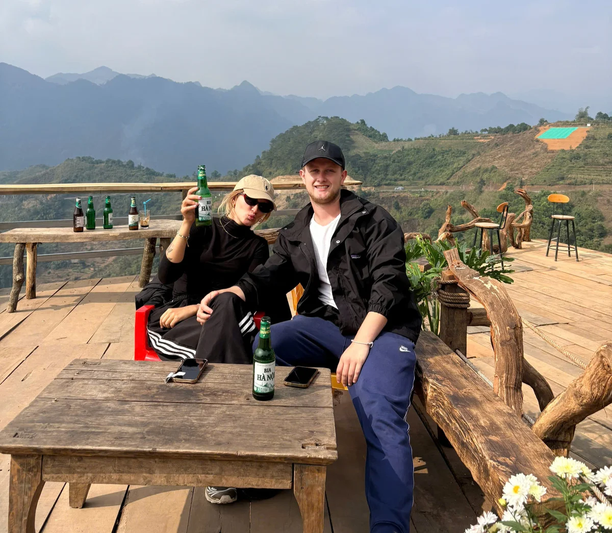 Two people smiling, sitting on a wooden deck with mountain views, holding beer bottles. Casual attire, sunny day, relaxed mood.