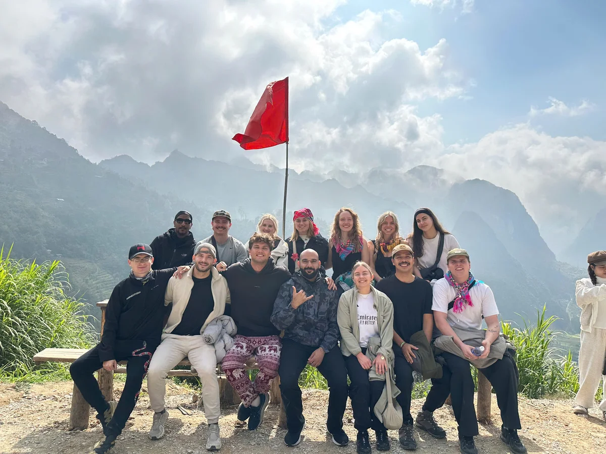 Group of smiling people posing on a mountain path with a red flag, under a cloudy sky. Lush green hills in the background.