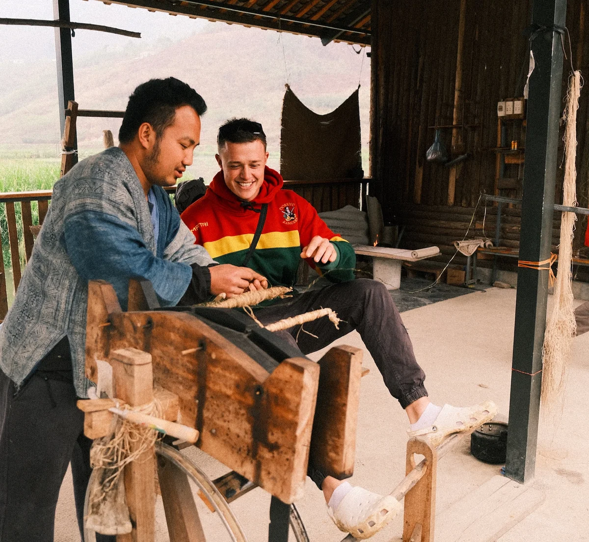 Two men are weaving yarn, one in a colorful hoodie. They sit in a rustic setting, smiling, amidst wooden tools and natural fibers.