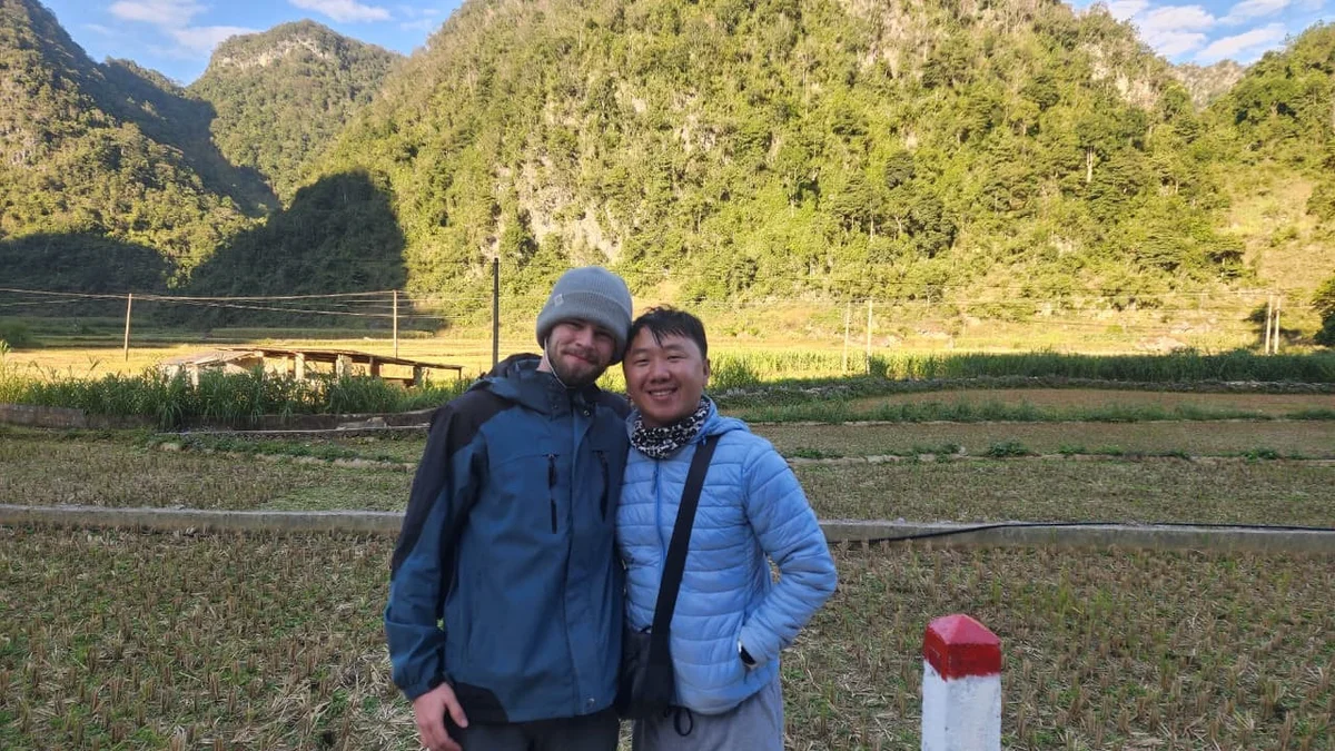 Two people in jackets smile, standing in a lush green field with mountains in the background. The sky is clear, creating a joyful atmosphere.