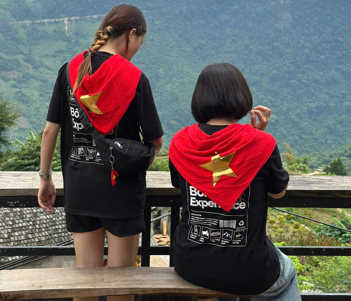 Two people with red scarves featuring yellow stars, facing away, overlooking a lush green valley. Text on black shirts reads "Bố Experience."
