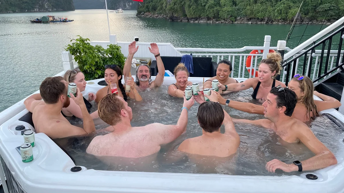 Group of people in a hot tub toasting with drinks, surrounded by water and greenery. Joyful mood with laughter and raised glasses.