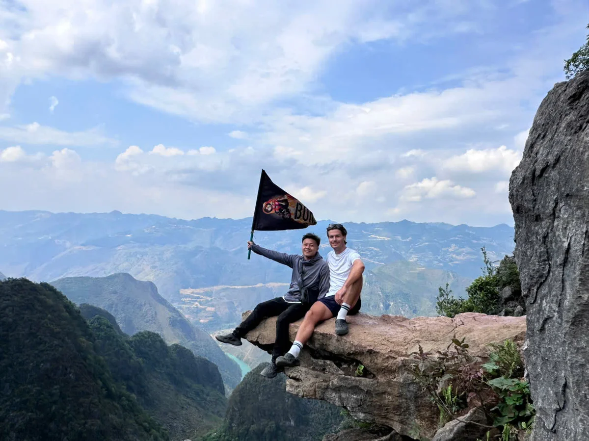 Two people pose on a cliff edge one waves the Bong Hostel flag