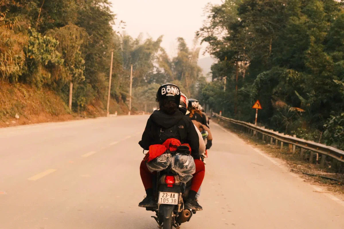 Two people ride a motorcycle on a rural road with trees. Helmet text reads "BỐNG." Road signs and license plate visible. Overcast mood.