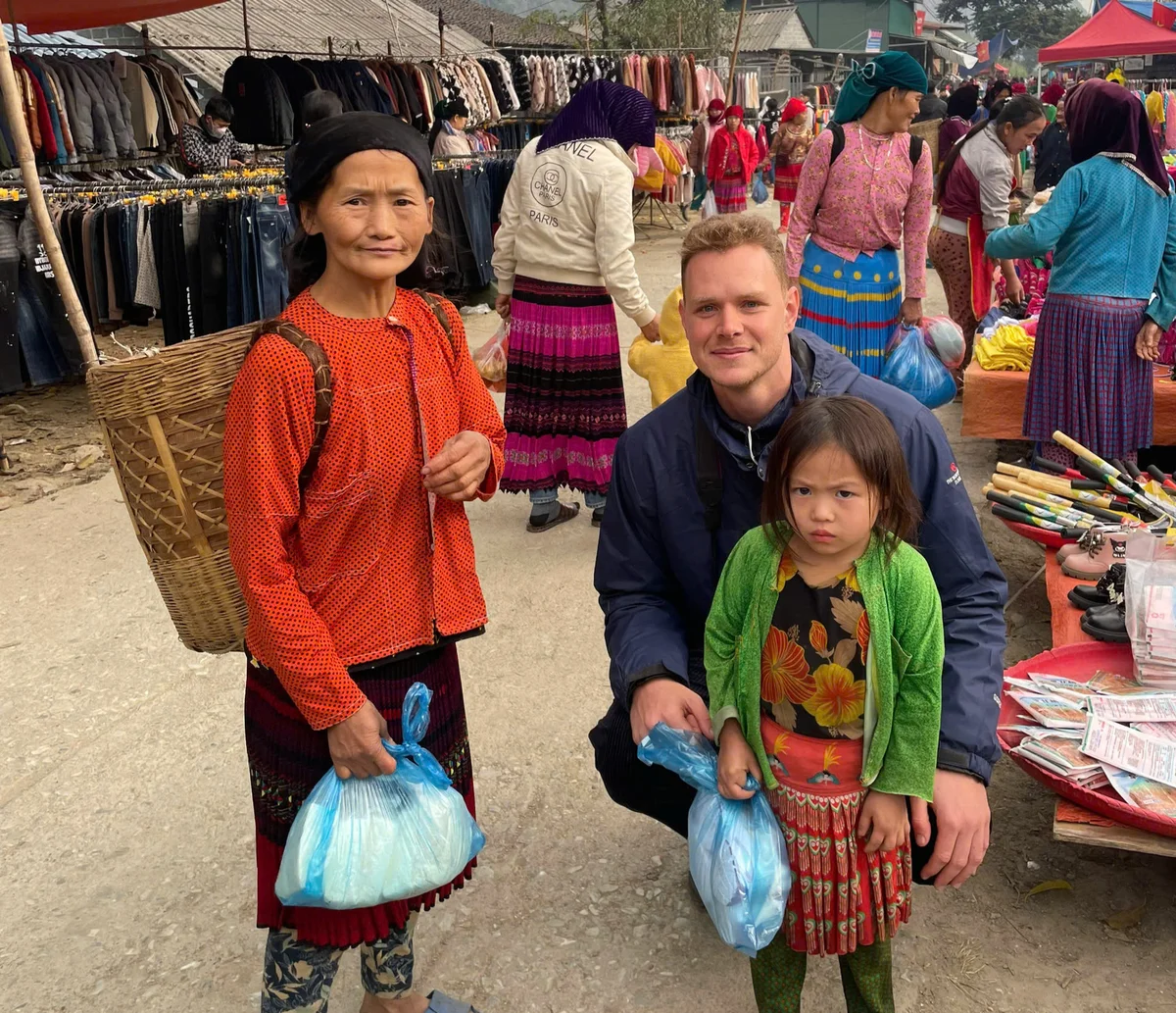A woman and child hold bags at a vibrant outdoor market. The woman wears orange and a wicker basket; the child wears green. Colorful stalls surround them.