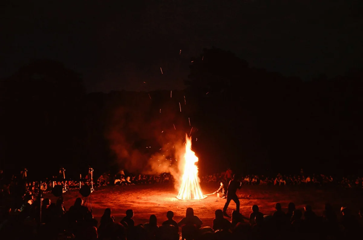 Crowd gathers around a large bonfire at night in a dark, open field. The fire illuminates the scene, creating a warm, lively atmosphere.