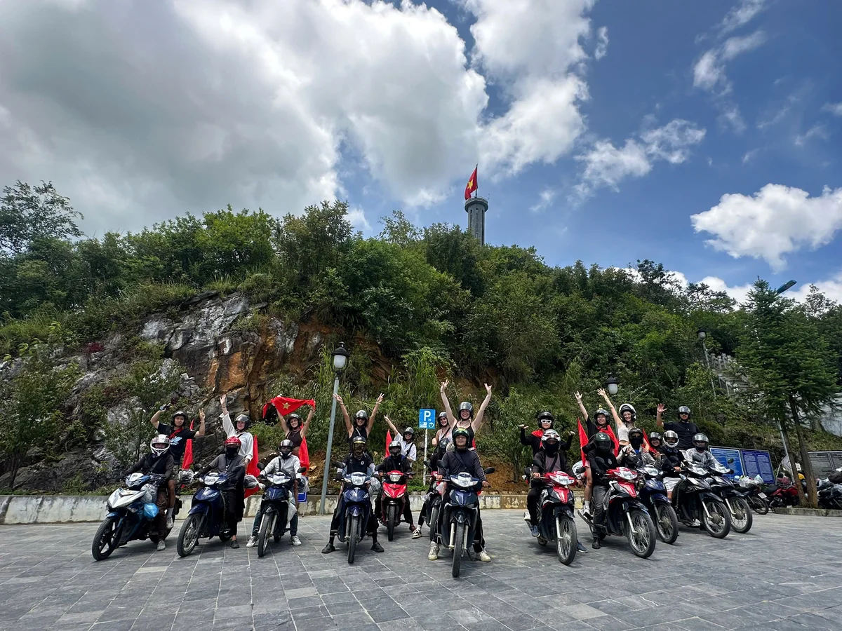 Group of people on motorcycles with flags, posing excitedly near a mountainous backdrop. The sky is cloudy, with a tower and flag visible.