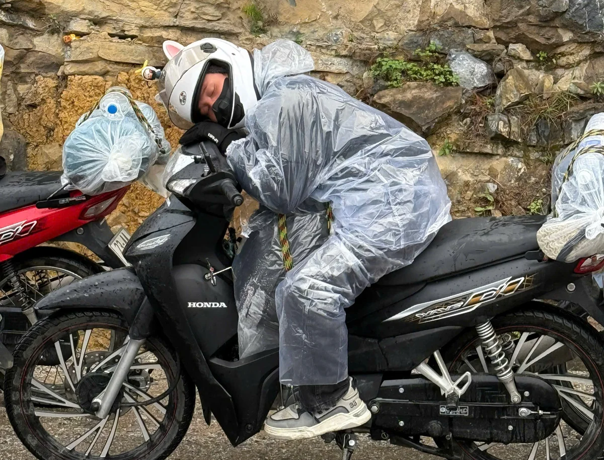 Person in a white helmet and rain poncho sleeps on a black Honda scooter, parked on a wet roadside with rocky background.