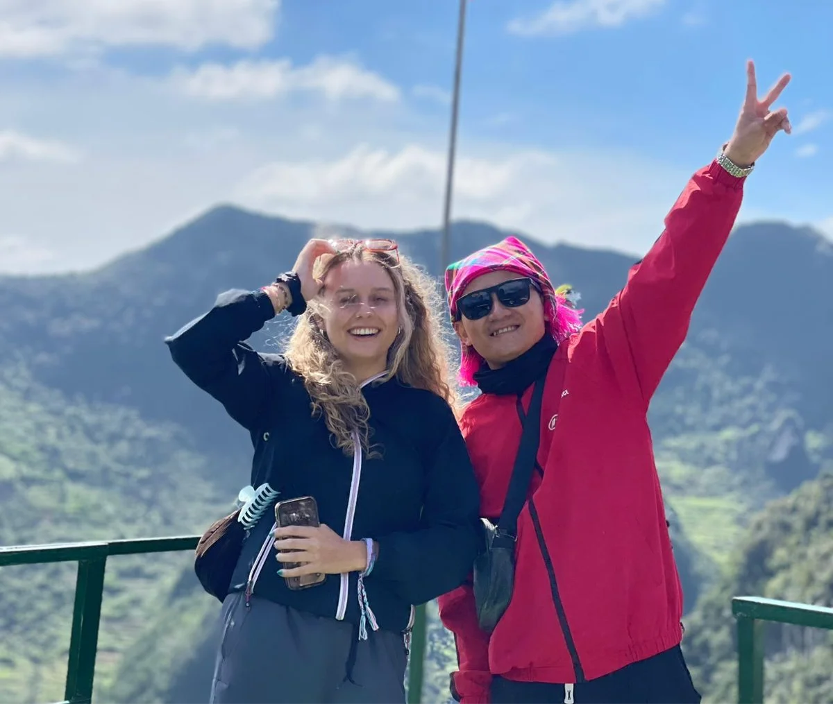 Two people smiling on a scenic mountain overlook. One wears a black jacket, the other a red jacket and pink headscarf, flashing a peace sign.