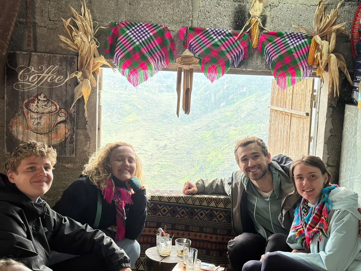 Group of four smiling people sit in a cozy room with patterned decor and green view. "Coffee" sign on wall. Bright scarves, cheerful mood.