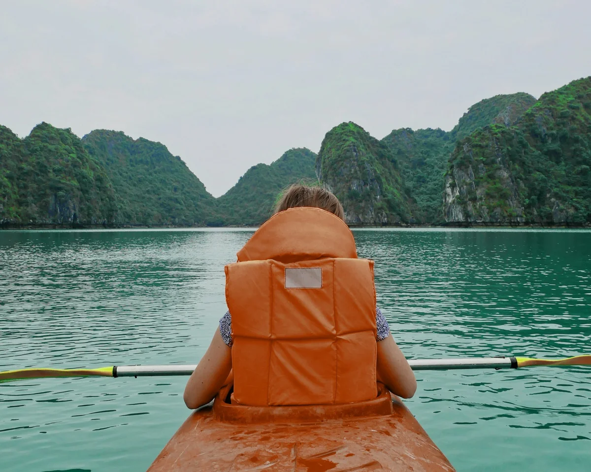 Person kayaking in calm turquoise water, wearing an orange life vest. Lush green hills form a serene, overcast background.