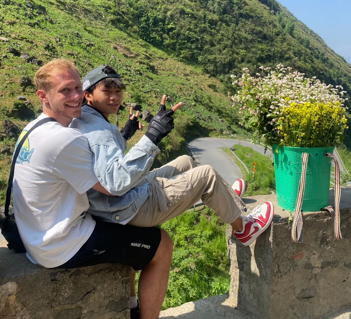 Two people smiling on a stone wall, one making peace signs, beside a basket of flowers. Green hillside and winding road in background.