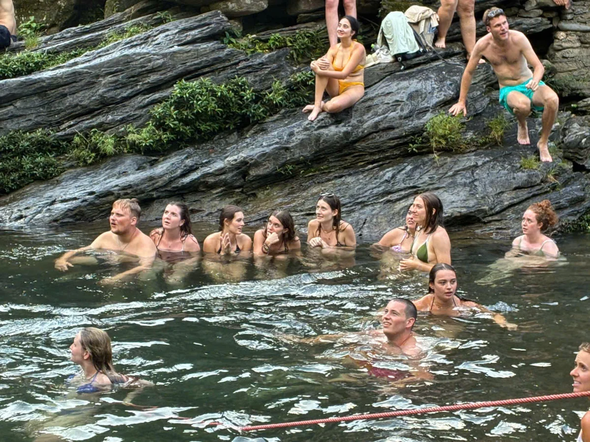 People swimming and relaxing in a rocky, natural pool setting. Some are seated on rocks, while others swim, creating a lively, joyful scene.