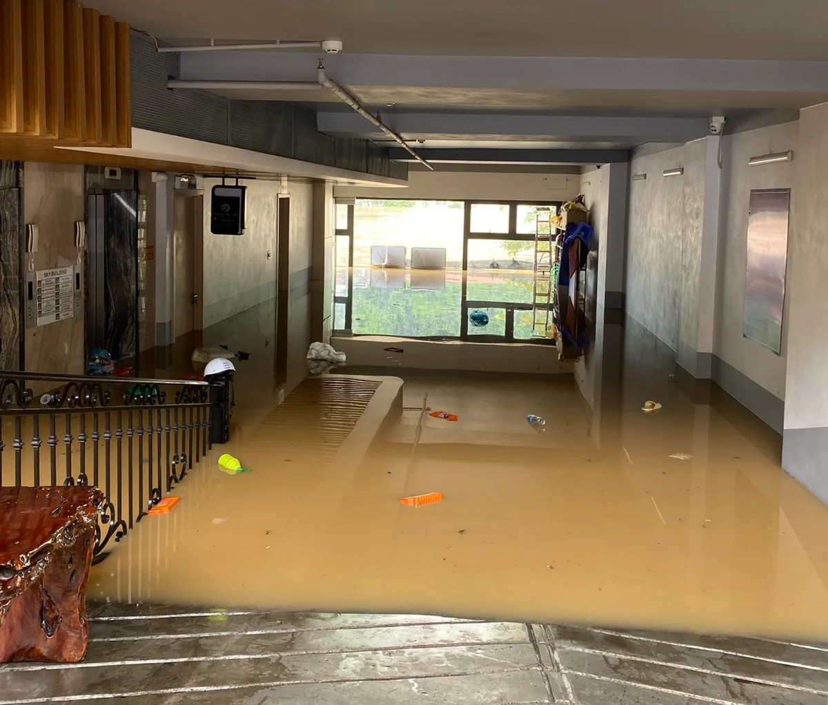 Flooded building lobby with muddy water covering the floor, debris floating. Sunlit window in the background, somber mood.