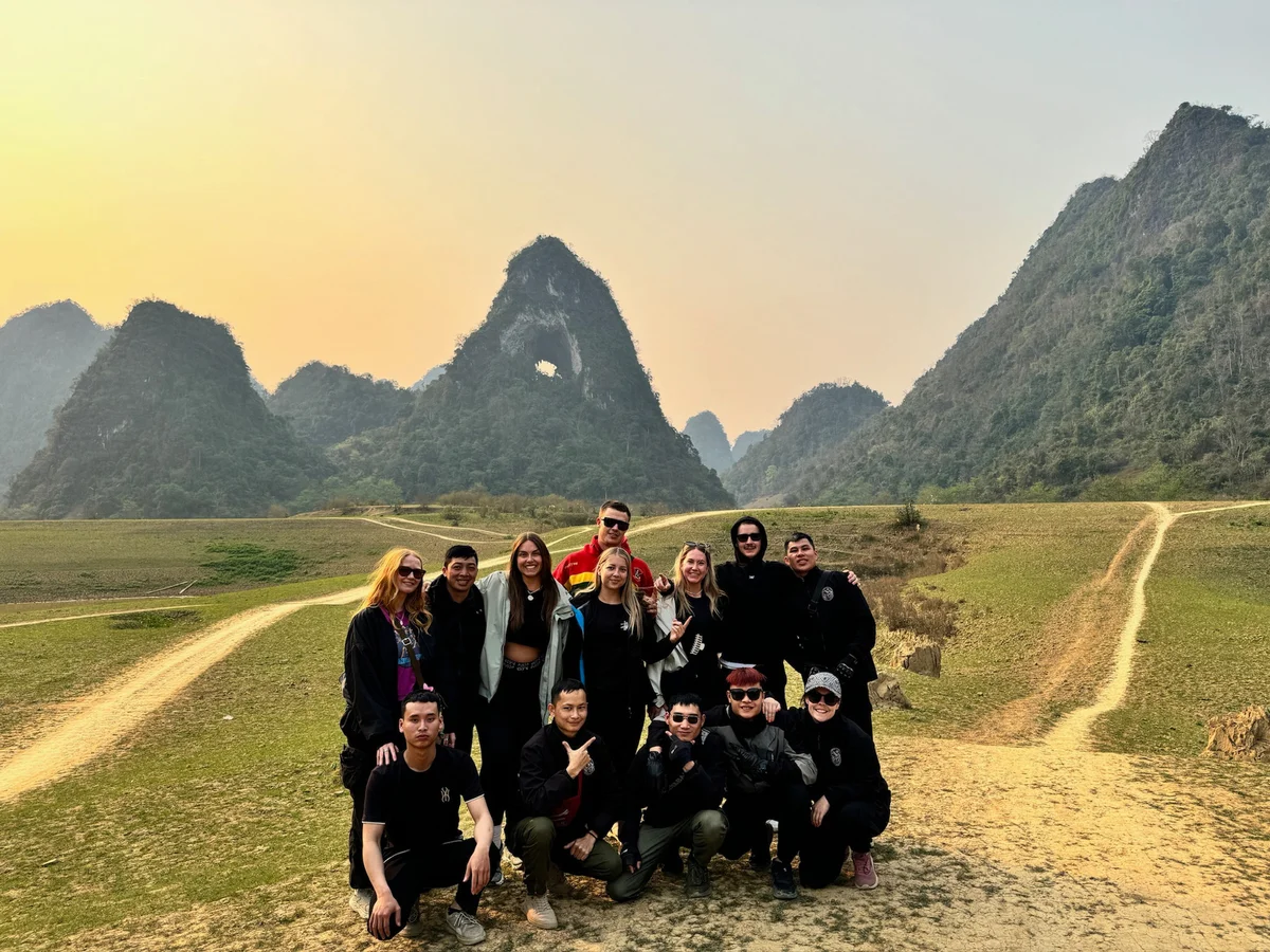 Group of people posing outdoors in a mountainous landscape with green hills and a hazy sky. Some are smiling, creating a joyful mood.