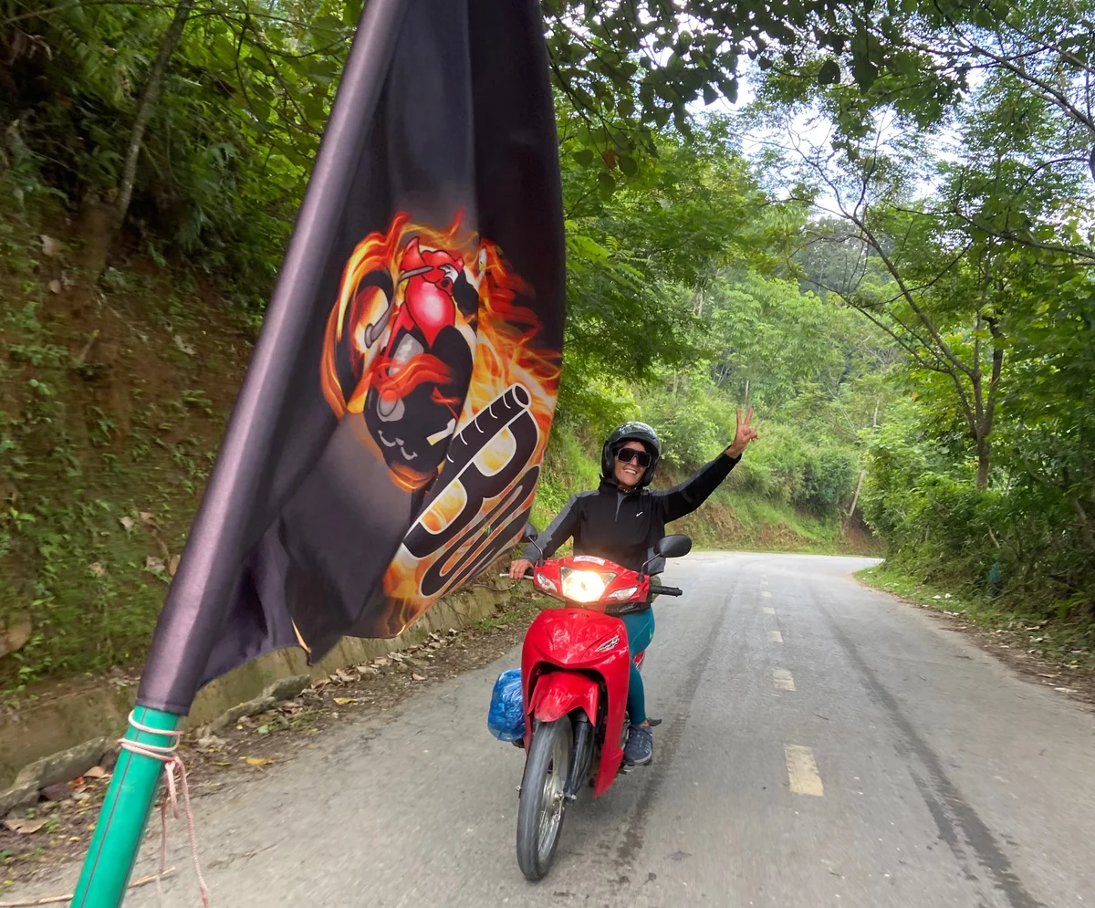 A motorbiker puts the peace symbol up to the camera with his fingers, there is a bong hostel flag in the shot