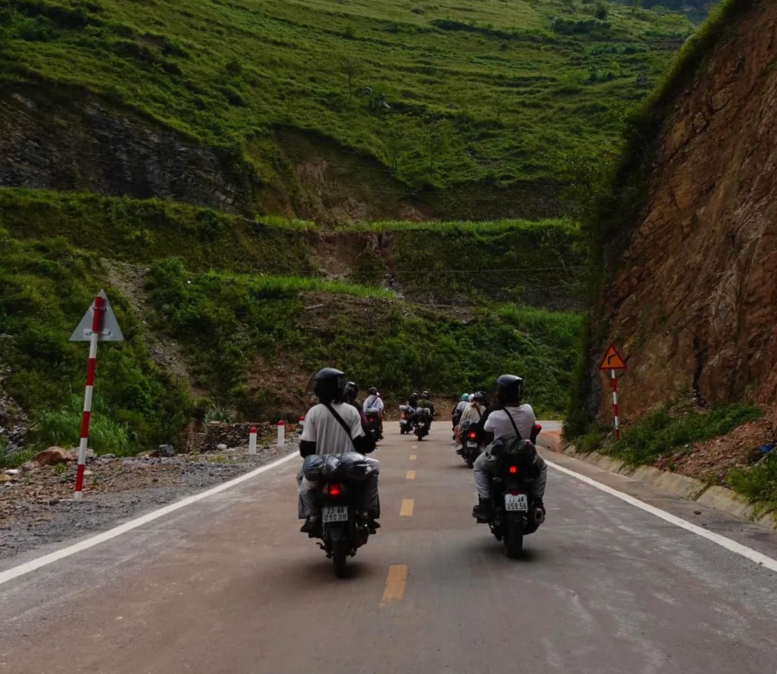 Motorcyclists ride through a scenic mountain road with lush green hills. Road signs and helmets are visible. A calm, adventurous mood.