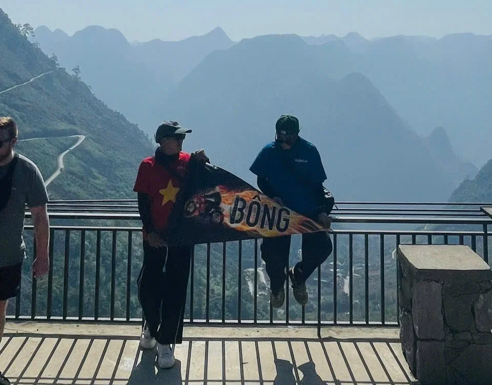 Two people hold a "BONG" sign on a scenic mountain overlook. One wears a red shirt with a star, the other blue. Curved road and mountains behind.