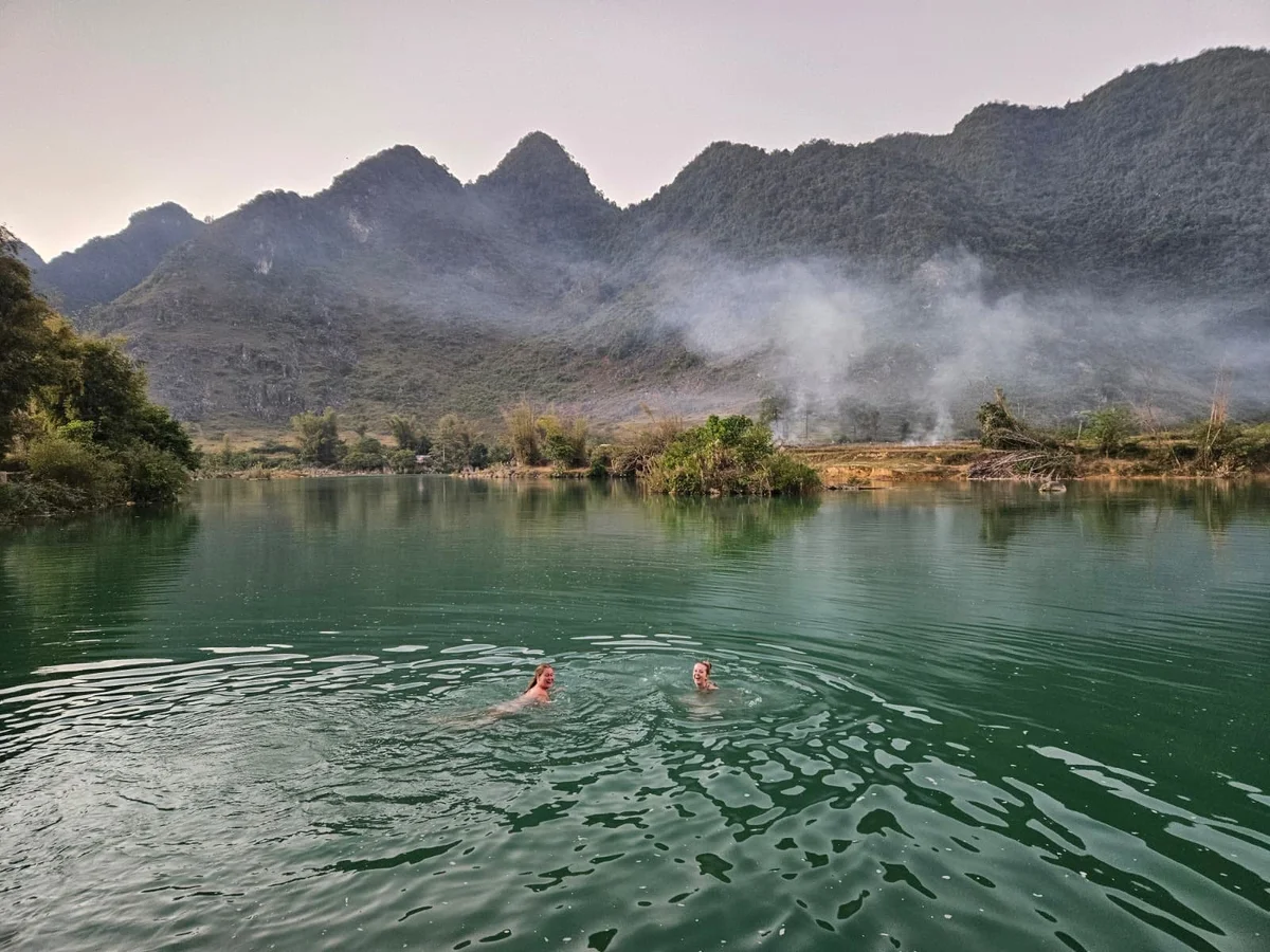 Two people swimming in a green lake with misty mountains in the background. Smoke rises from the shore, creating a serene mood.