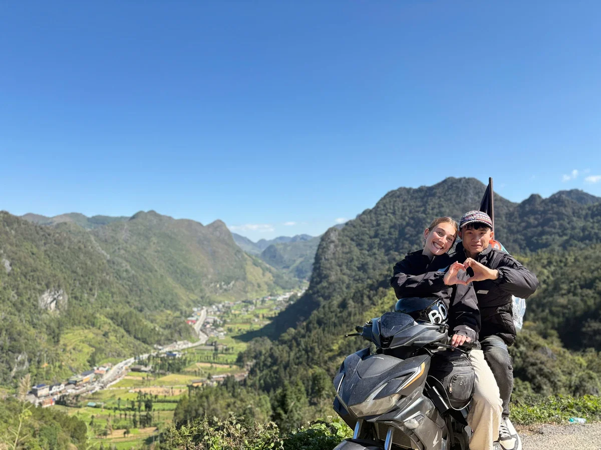 Two people on a motorcycle make a heart shape with their hands, smiling. They are in a mountainous landscape under a clear blue sky.