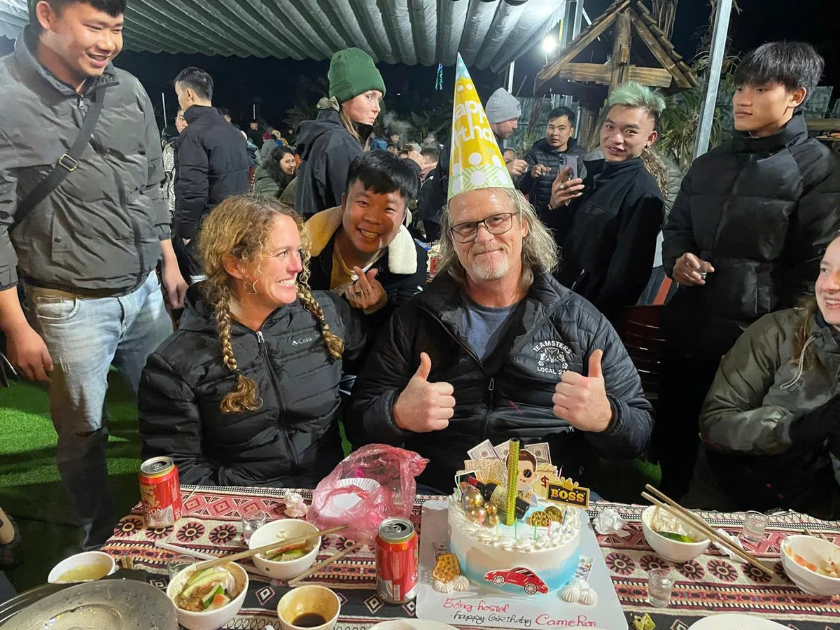 Man with hat and glasses gives thumbs up at outdoor birthday party. Cake with car design on table; guests smile around him under nighttime tent.