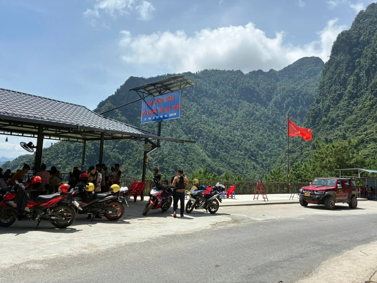 Motorbikes parked at a roadside cafe with people in helmets. Red flag and vehicle nearby. Lush mountains in the sunny background. Sign overhead.