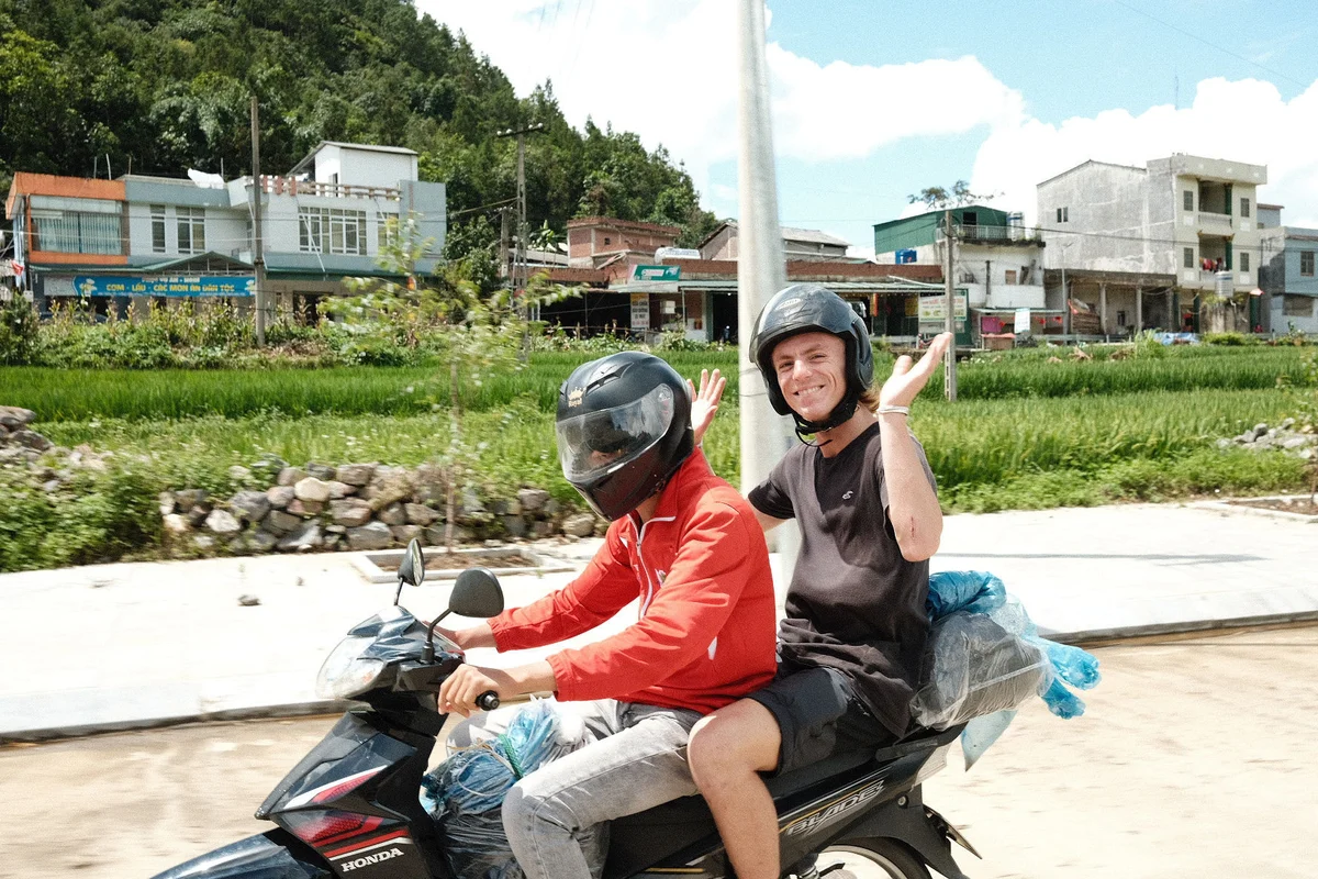 Two people on a motorbike; the driver in red, the passenger smiling with arms raised. Rural setting with a lush green backdrop and buildings.